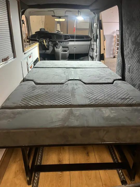 Rozłożone łóżko w VW T6 z pikowaną tapicerką, widok w stronę kabiny