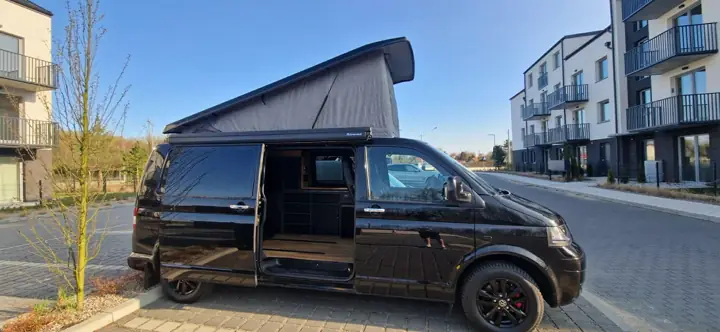 Czarny VW T6 z rozłożonym dachem i otwartymi drzwiami bocznymi na osiedlu